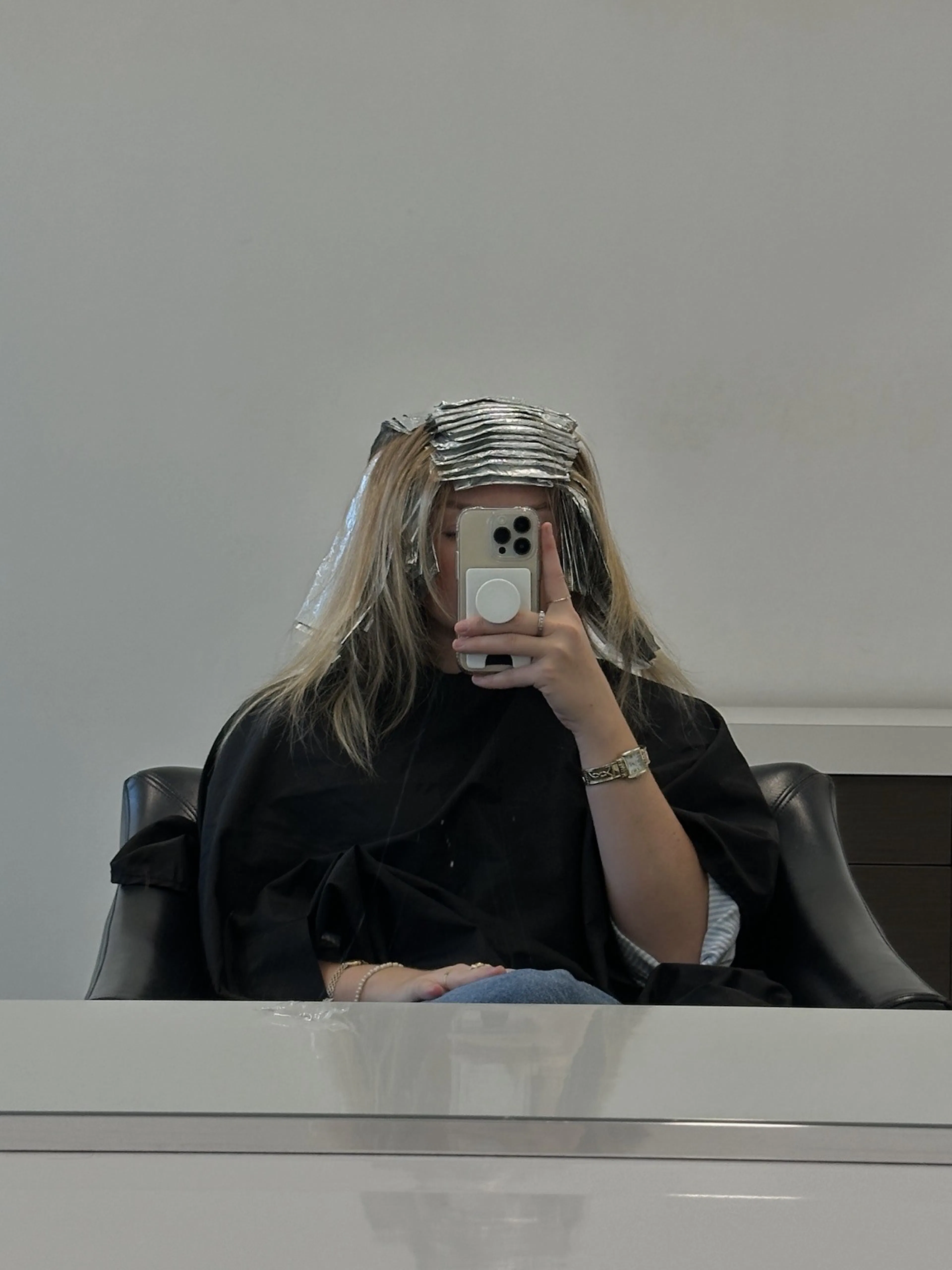 A client in a salon chair with foils in her hair, taking a mirror selfie.