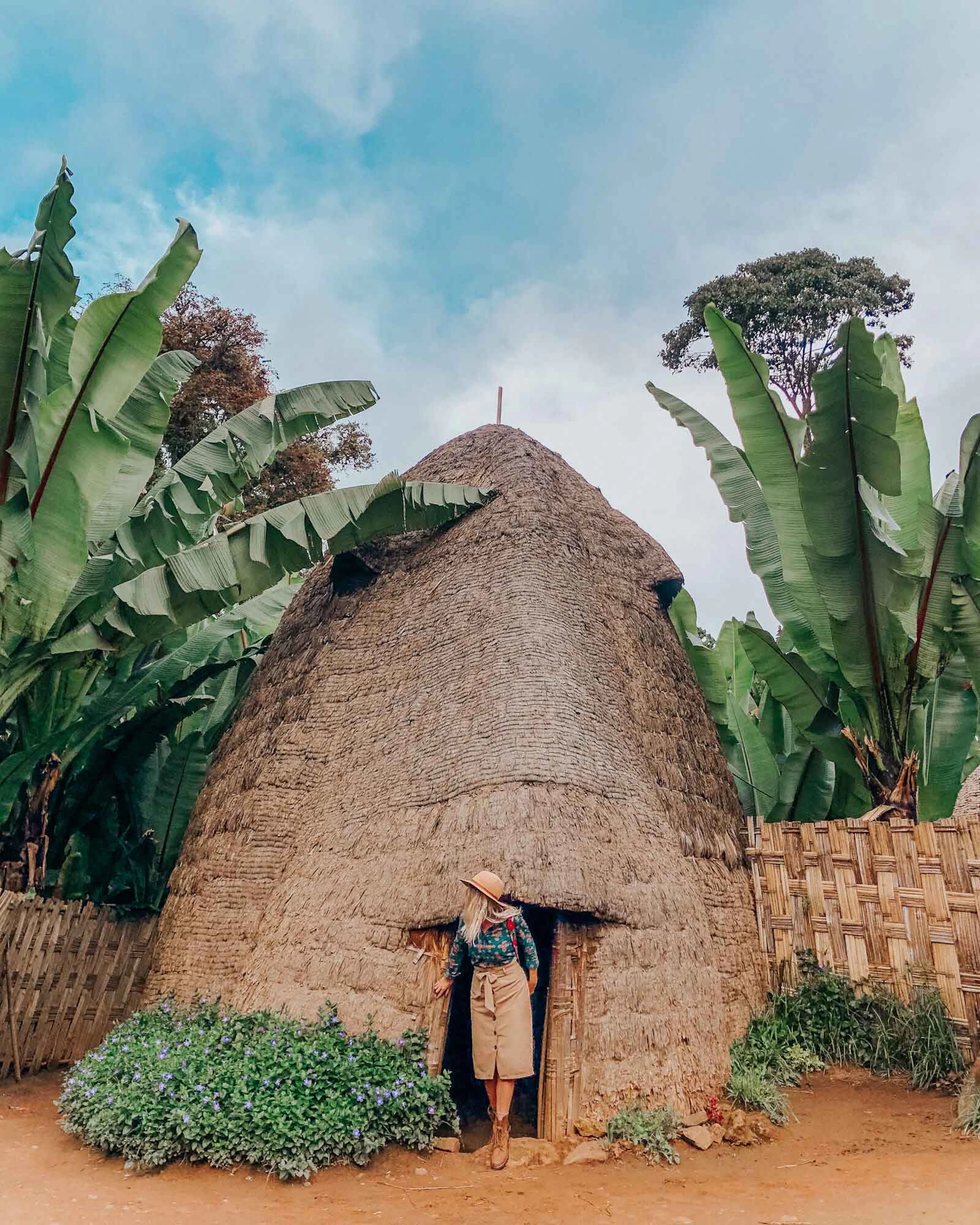 A person in the doorway of a tall thatched hut among banana trees