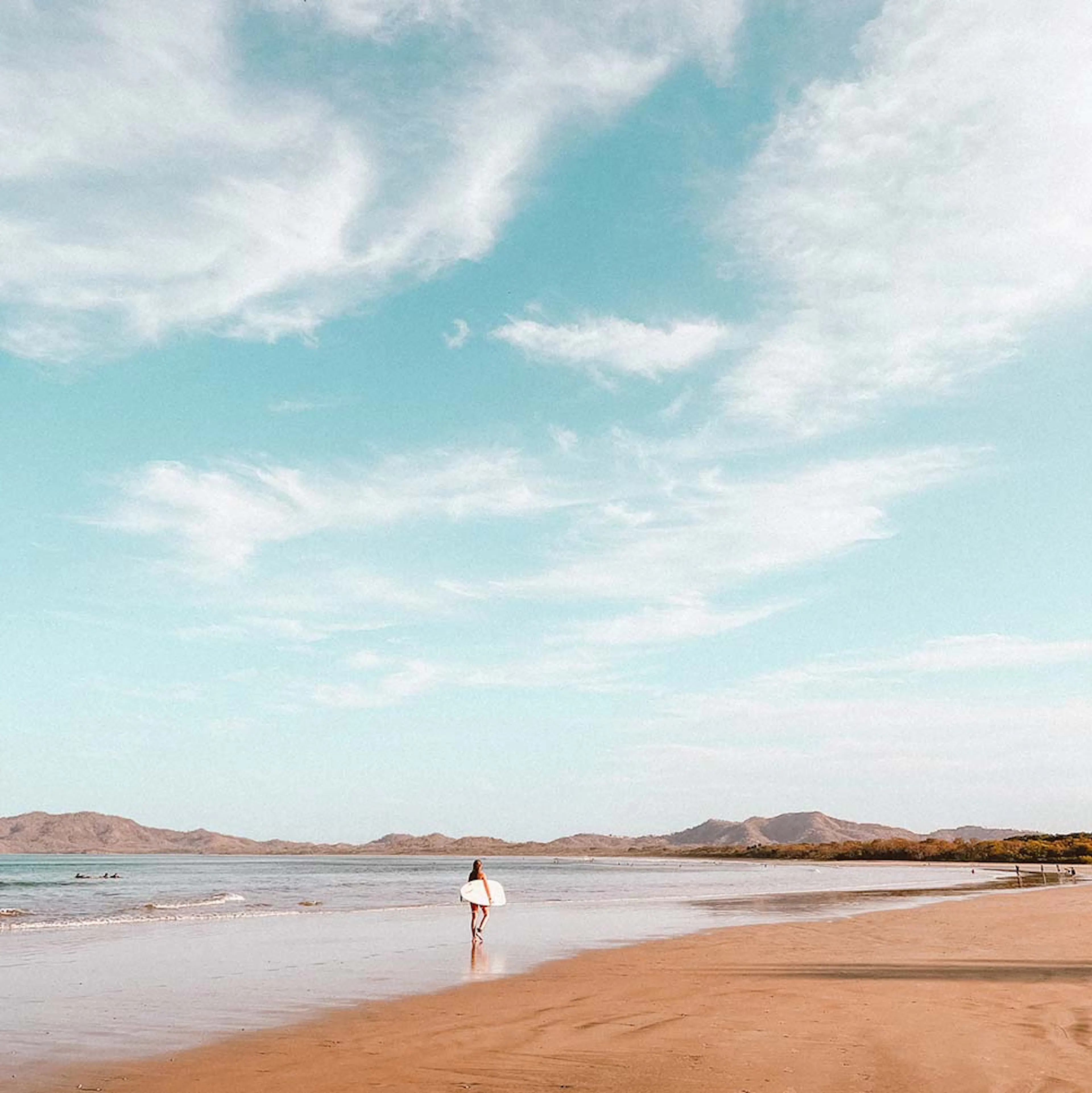 A surfer walking along a wide beach under a blue sky
