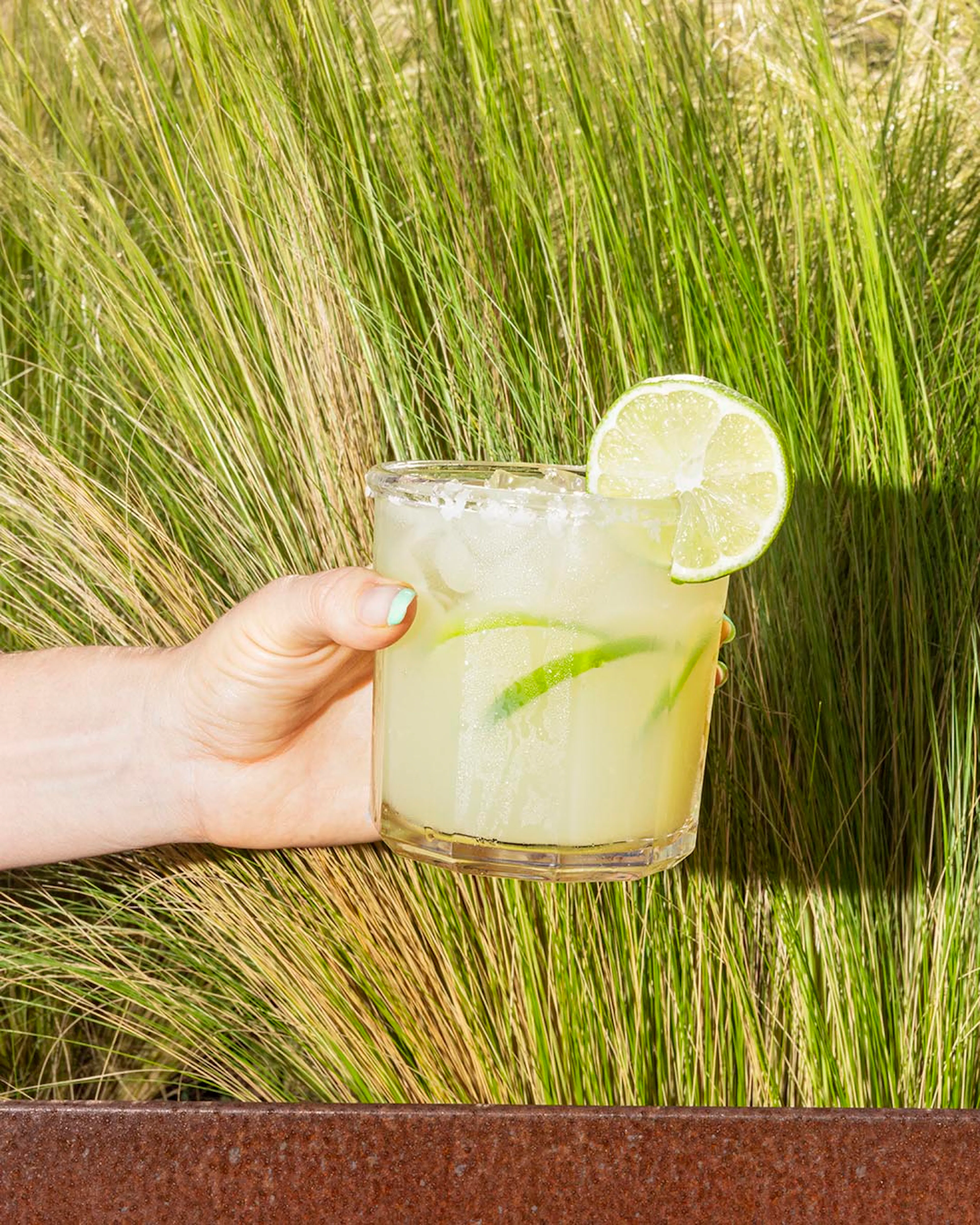 A hand holding a margarita with a lime wedge in front of tall grass