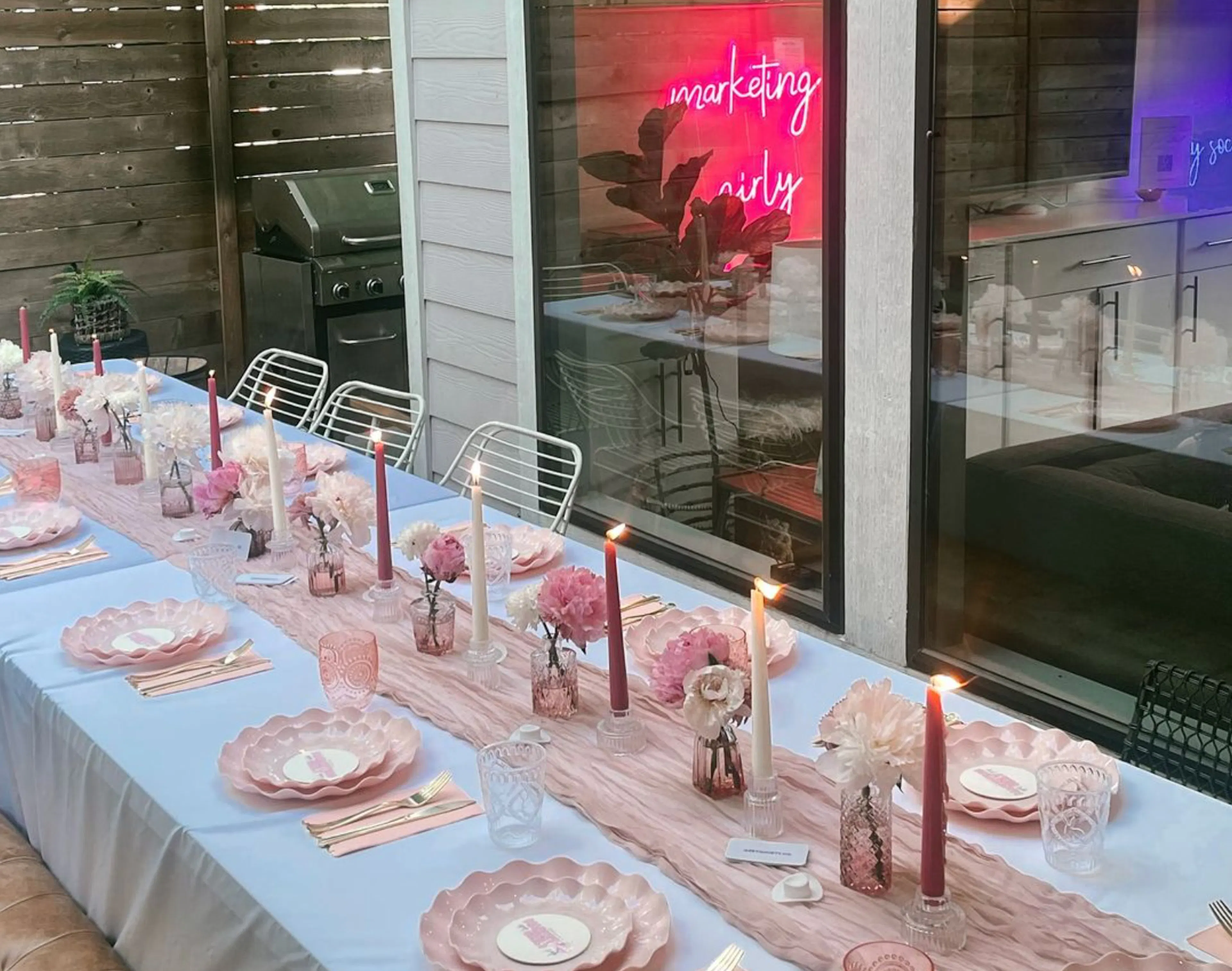 A long dinner table set with pink plates and candles under a “marketing girly” neon sign