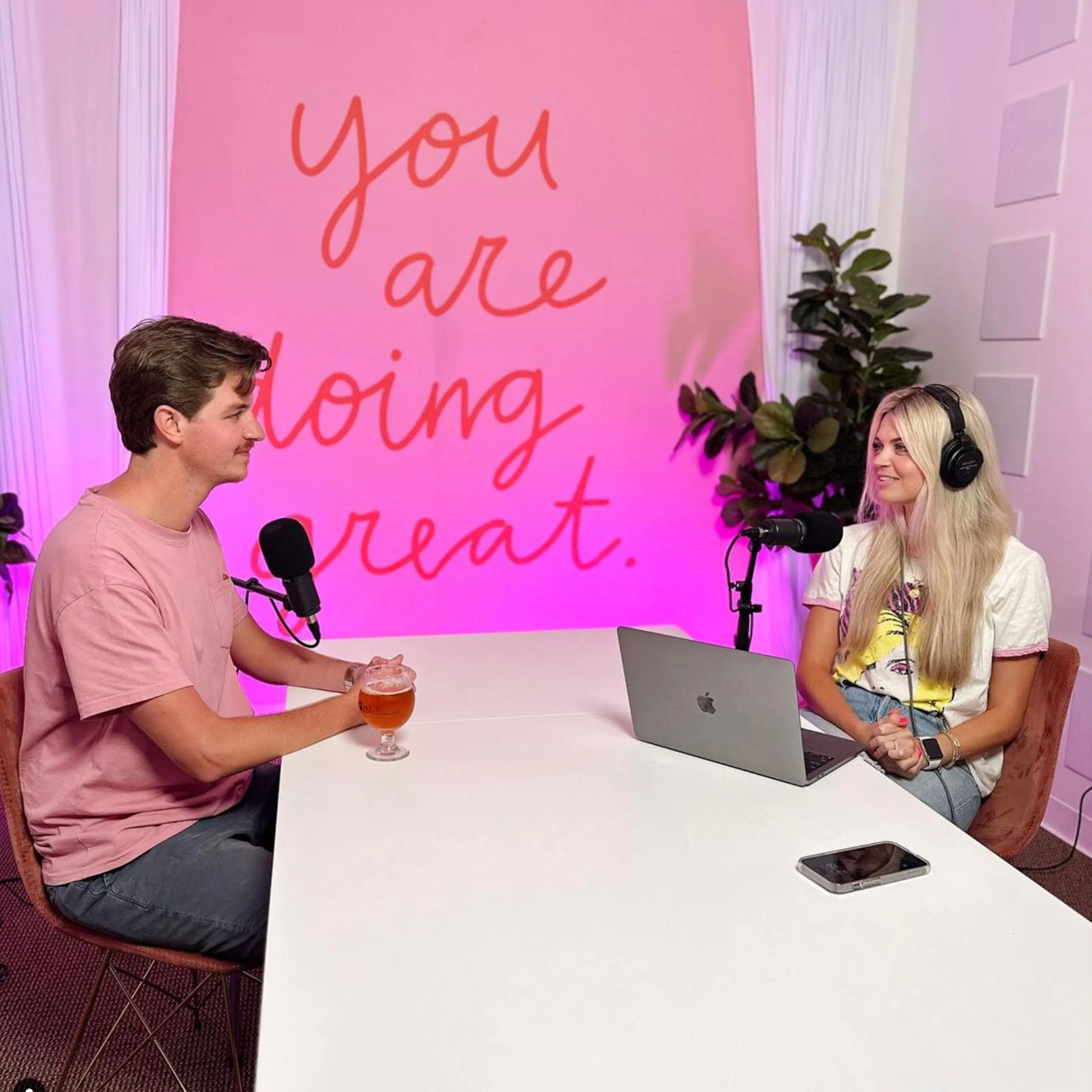 Two podcast hosts talking at a table under a pink “you are doing great” sign