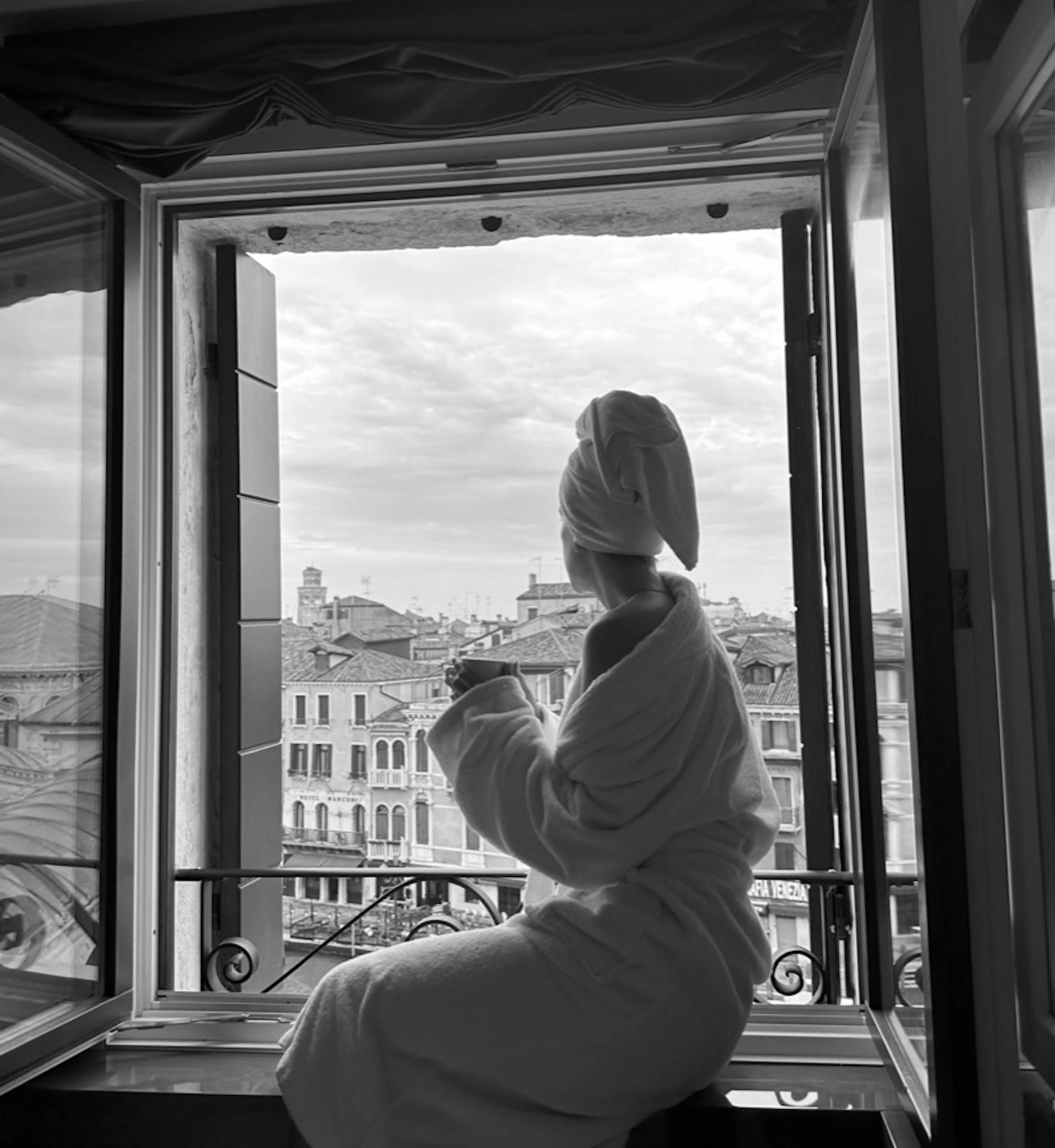 A black-and-white photo of a woman in a robe and towel turban sitting in an open window, looking out over the rooftops.